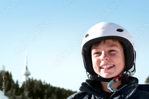 Winter ski vacation in mountains resort, alpine skiing concept. Smiling happy child skier in ski helmet against background of mountain forest and blue sky. Close-up portrait, Banner, copy space © Elena