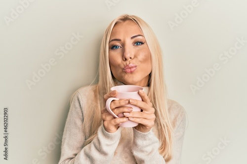Young blonde woman drinking a cup of coffee looking at the camera blowing a kiss being lovely and sexy. love expression.