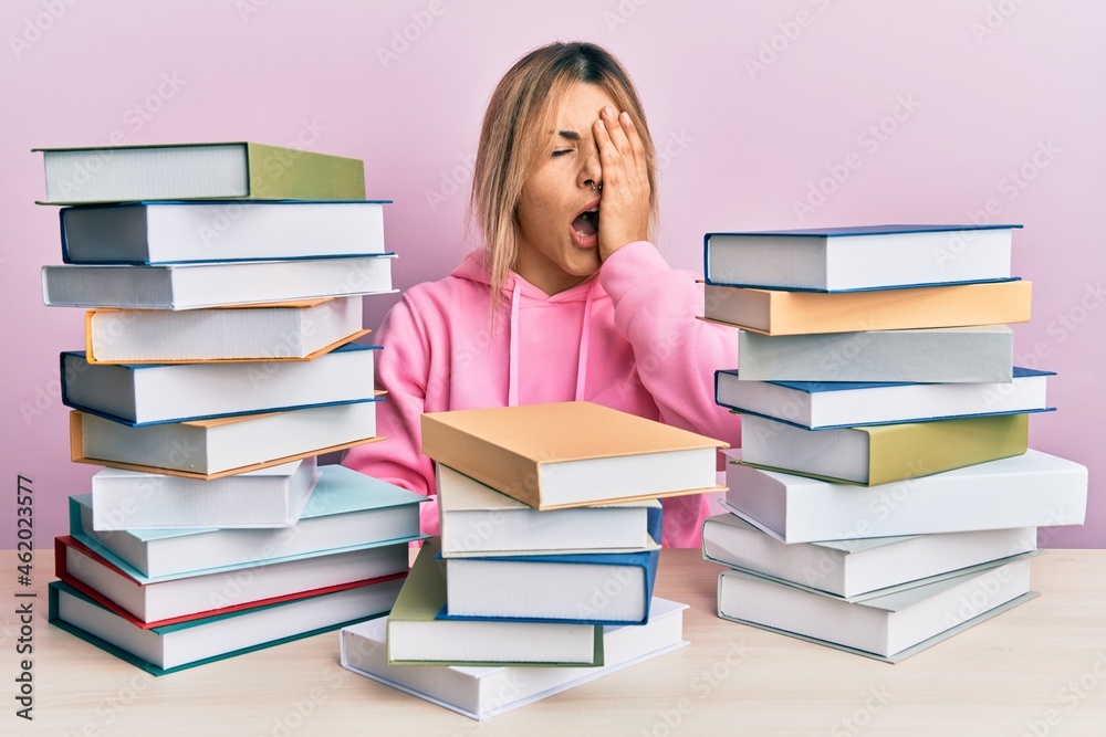 Young caucasian woman sitting on the table with books yawning tired covering half face, eye and mouth with hand. face hurts in pain. Young caucasian woman sitting on the table with books yawning tired covering half face, eye and mouth with hand. face hurts in pain.