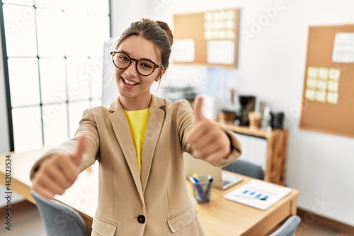 Young brunette teenager wearing business style at office approving doing positive gesture with hand, thumbs up smiling and happy for success. winner gesture.