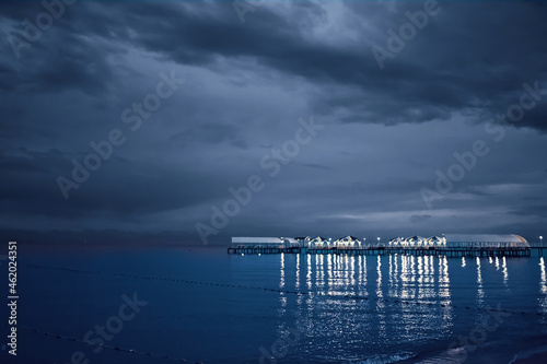 Twilight landscape of pier stretching out into sea. Cloudy sky. Illumination and umbrellas on ocean bridge. Nature summer landscape. Tourism background. Travel and vacation. Glare from lamps on water.
