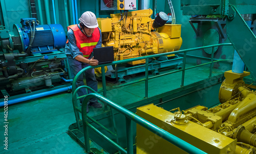 Asian young engineer in reflective clothing with helmet using laptop computer to checking engine quality system inside of engine room of fishing vessel photo