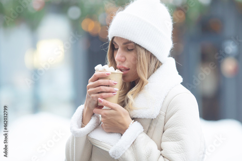 Smiling beautiful girl in knitted hat drinking hot cocoa with marshmallows.