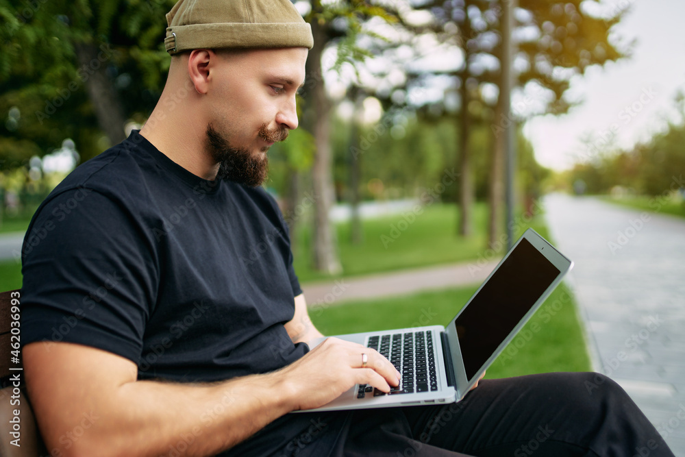 Freelancer sits in the city park while typing on laptop. Blogger hipster traveler Young stylish man