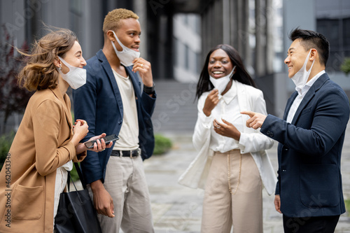 Multiracial business people taking off medical masks and talking on city street. Concept of health protection during Coronavirus pandemic. Joyful businessmen and businesswomen wearing formal wear