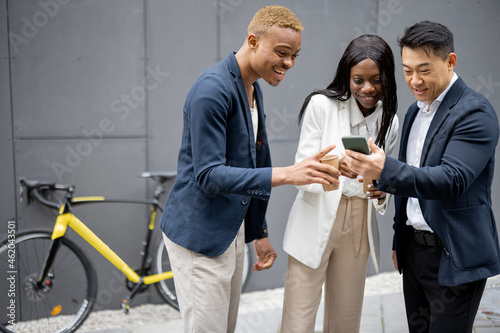 Multiracial business team watching something on smartphone on city street. Concept of break on job. Idea of team building. Smiling businesswoman and businessmen wearing formal wear