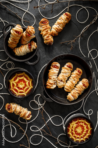 Mummy dogs made of sausages and puff pastry with addition of ketchup-mustard sauces on a black background top view. Halloween food idea for party 