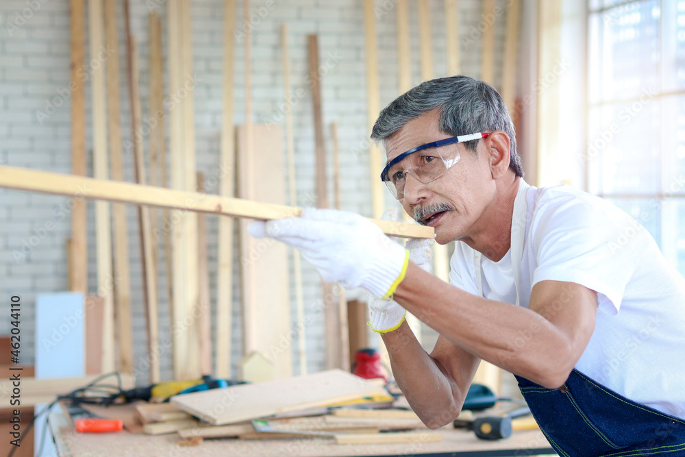 Elderly carpenter wearing safety glasses goggles and cloth gloves, holding and aiming wooden plank, senior craftsman estimating length of timber by his eyes at carpentry workshop, enjoy DIY hobby. Elderly carpenter wearing safety glasses goggles and cloth gloves, holding and aiming wooden plank, senior craftsman estimating length of timber by his eyes at carpentry workshop, enjoy DIY hobby.
