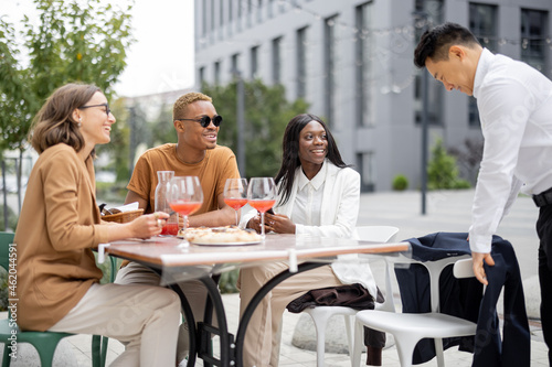 Happy multiracial business team having lunch with wine and pizza at outdoor cafe. Concept of teambuilding and corporate event. Idea of rest and leisure on job. Asian man came to his colleagues