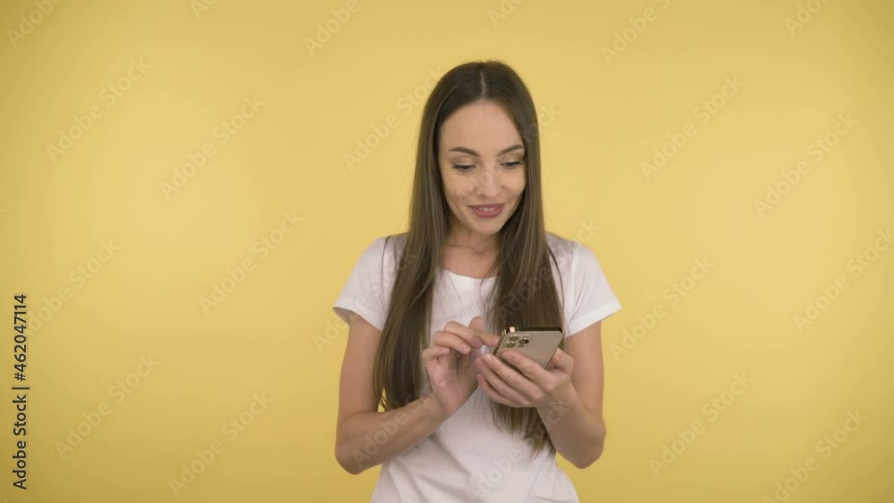 Smiling long-haired woman middle age with a smartphone having thumbs up on yellow background in studio. Middleweight woman in basic white t-shirt Smiling long-haired woman middle age with a smartphone having thumbs up on yellow background in studio. Middleweight woman in basic white t-shirt