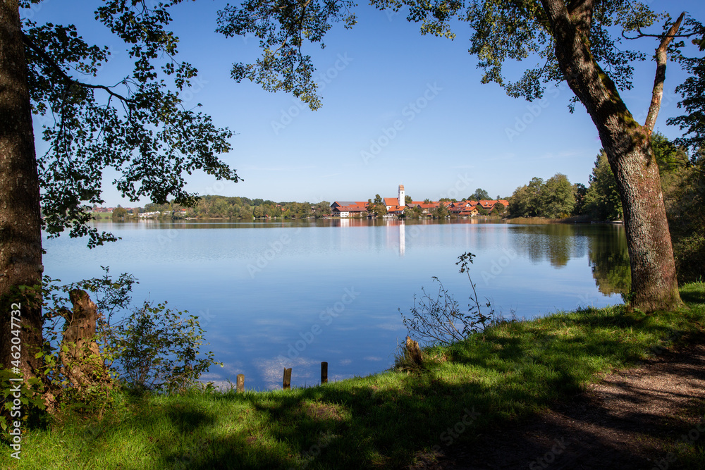 View over an idyllic lake in Bavaria with reflective village including white church tower. In the foreground trees with shadows at a bathing area