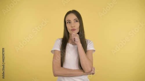 Portrait of a cute attractive woman 40 years old on an isolated yellow background in the studio. Long-haired woman in basic white t-shirt meditates over the proposal of the touching chin