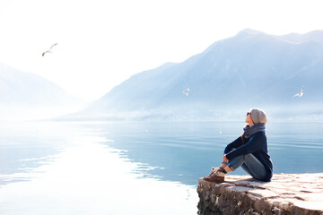 Young woman relaxing at winter sea beach. Traveler resting by blue mountain