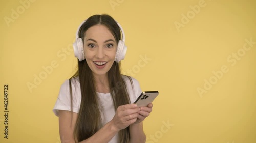 Funny attractive woman 35 years old middle age on yellow background in studio in profile. Woman having fun dancing listening music in big white wireless headphones holding smartphone in her hand
