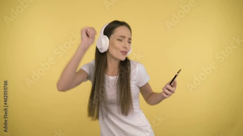 Funny attractive woman 35 years old middle age on yellow background in studio in profile. Woman having fun dancing listening music in big white wireless headphones holding smartphone in her hand