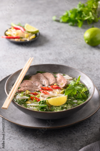 Pho Bo Soup with beef, rice noodles, lime, chili pepper in gray bowl on gray backgound. Vietnamese cuisine. Vertical format.