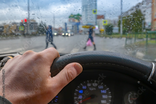view of the driver hand on the steering wheel of a car against the background of a wet windshield with raindrops and people walking along a pedestrian crossing at an intersection with a traffic light