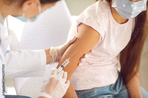 Kid getting a flu shot at a modern clinic or hospital. Doctor giving an injection in the arm to a child in a face mask. Nurse gives a new effective Covid 19 vaccine to a little girl  close up