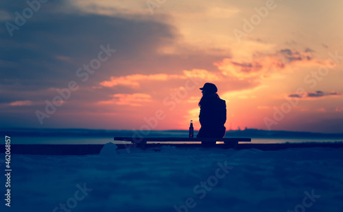 A young lonely girl sits on a bench and is sad next to a bottle of beer. Female alcoholism. Depression and loneliness, alcohol concept. Sunset and evening winter silhouette.