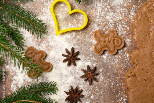 Two unbaked gingerbread man cookies, three star anise fruits, a cookie cutter, fir branches and a piece of rolled out dough are lying on a surface sprinkled with flour.