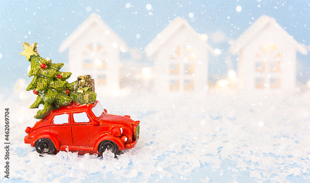 Christmas decor - red retro car on snow carries past white houses with lights garlands in bokeh Christmas tree with gift boxes on roof. Toy on blue background, space for text. New Year in town