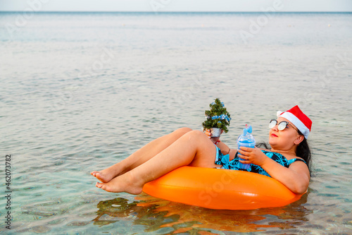 A woman in a swimming ring in a Santa Claus hat with a cocktail and a small Christmas tree in her hand floats in the sea during Christmas holidays.