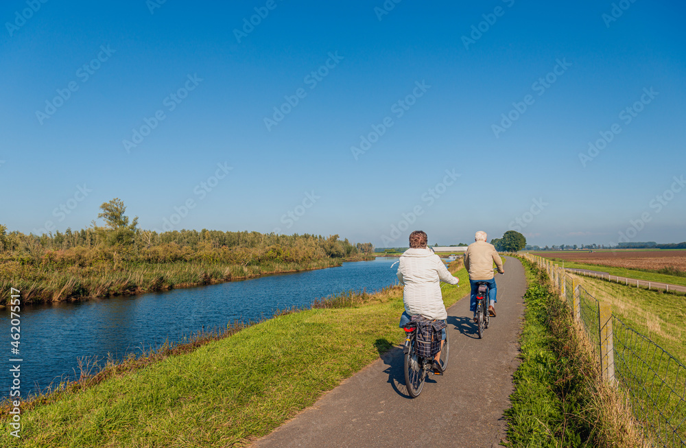 Elderly man and woman couple cycle one behind the other with their electric bicycles on a Dutch dike along the water of an old creek. It is a sunny day in the autumn season.