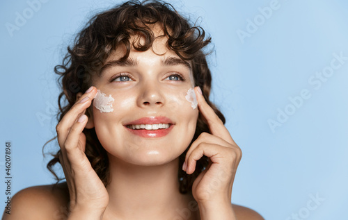 Beauty woman applying face cream on clean fresh skin, detoxifying effect, moisturize and hydrate facial with scrub, nourishing effect of mask, looking aside, blue background
