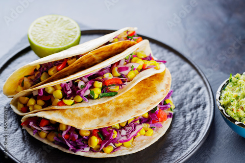 Vegetarian tacos with various vegetables, guacamole and lime on dark background. Tacos with sweet corn, purple cabbage and tomatoes on a black plate. Close-up