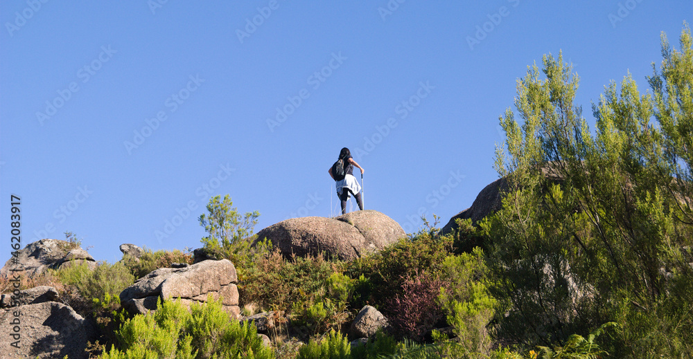 Mulher com equipamento de montanhismo no topo de uma pedra no cimo de uma montanha a observar a paisagem