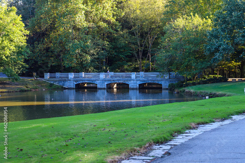 a stone bridge over the lake surrounded by green grass and lush green and autumn colored trees with still silky brown lake water at Lenox Park in Brookhaven Georgia USA photo