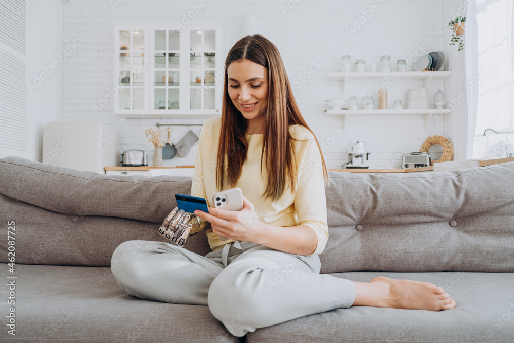 Pretty happy woman with prosthesis arm pays for online order with credit card via mobile phone on sofa in living room closeup Pretty happy woman with prosthesis arm pays for online order with credit card via mobile phone on sofa in living room closeup