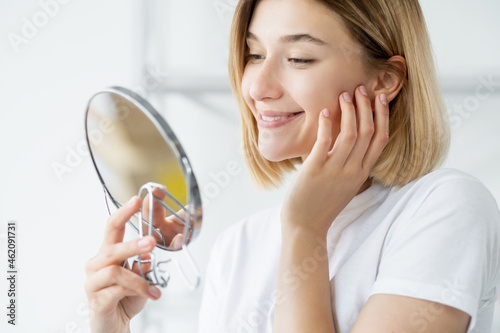 Morning skincare. Facial treatment. Skin rejuvenation. Satisfied smiling woman admiring radiant fresh face in mirror on defocused light background.