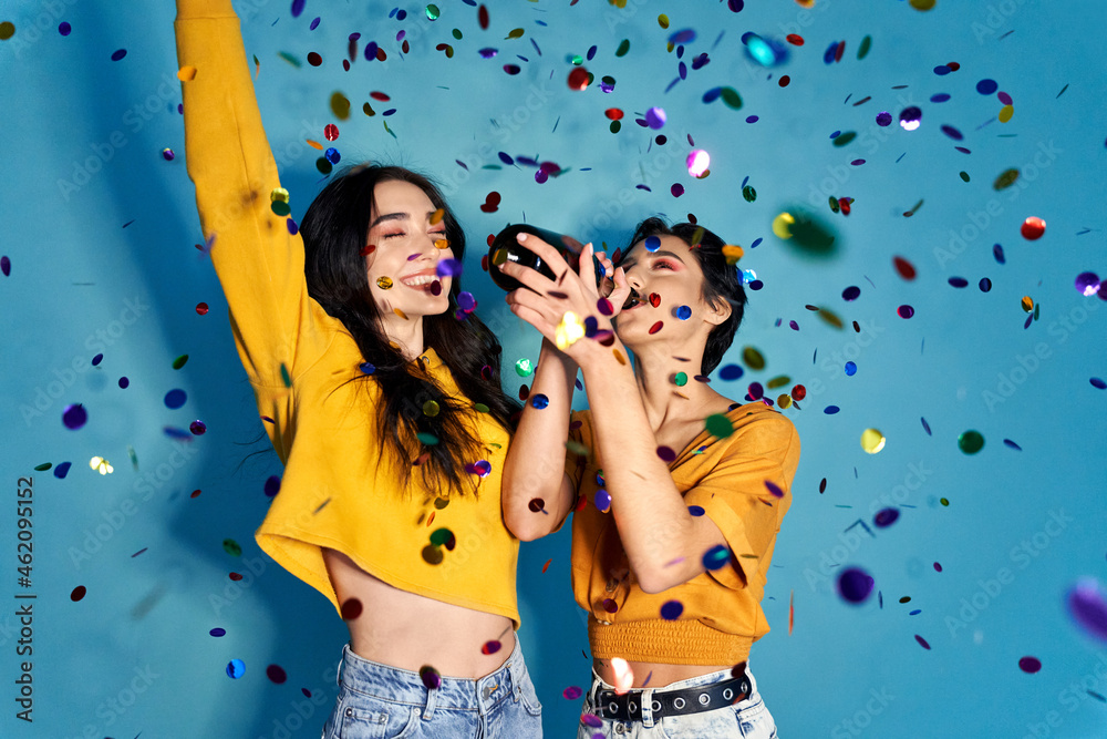 Two happy excited pretty stylish cool diverse gen z girls friends having fun celebrating New Year or birthday hen night club party event, dancing in confetti and drinking champagne on blue background. - obrazy, fototapety, plakaty 