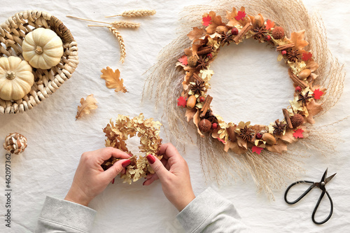 Hands making dried floral wreath from dry pampas grass and Autumn leaves. Flat lay on off white textile table cloth. Scissors, decorative pumpkins and dry oak leaves on the table. DIY Fall decorations