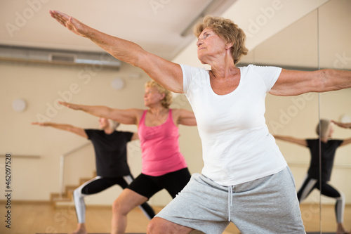 Concentrated senior woman performing standing lunging asana Virabhadrasana during yoga class with female group in fitness studio.