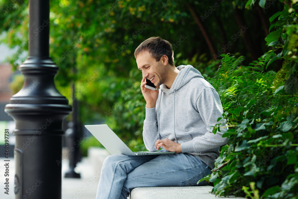 Happy man make phone call sit in park with laptop on knees working remotely from outside. Casual freelancer guy book hotel or plane tickets for distance work or business travel. Freelance occupation Happy man make phone call sit in park with laptop on knees working remotely from outside. Casual freelancer guy book hotel or plane tickets for distance work or business travel. Freelance occupation