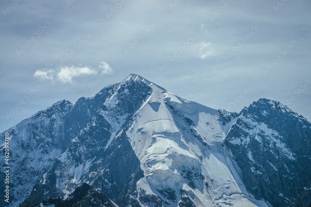 Awesome mountains landscape with big snowy mountain pinnacle in blue white colors and white cirrus clouds in blue sky. Atmospheric highland scenery with high mountain wall with pointed top with snow.
