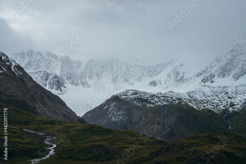Minimalist gloomy highlands landscape with snow-capped high mountains in overcast weather. Snowbound mountain range in low clouds. Atmospheric minimalism with green mountains, white snow and gray sky.