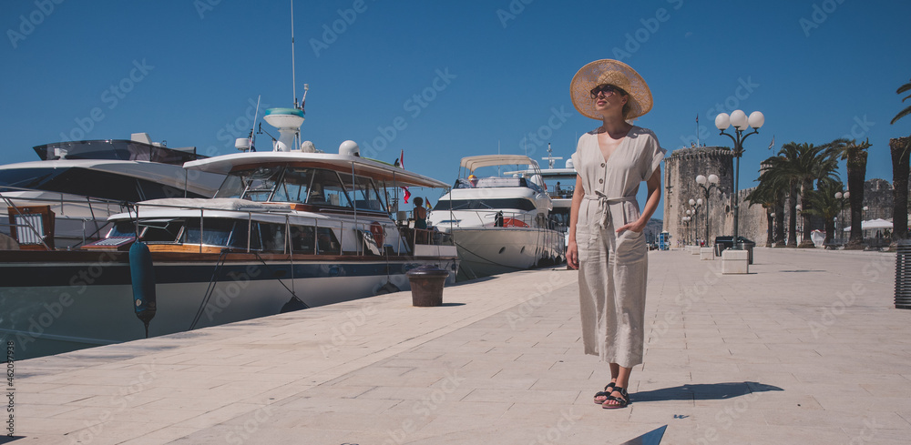 Young woman at the pier with sailing yachts. A beautiful fashionable girl walks near the moored sport yachts on a sunny hot summer day. Young woman at the pier with sailing yachts. A beautiful fashionable girl walks near the moored sport yachts on a sunny hot summer day.