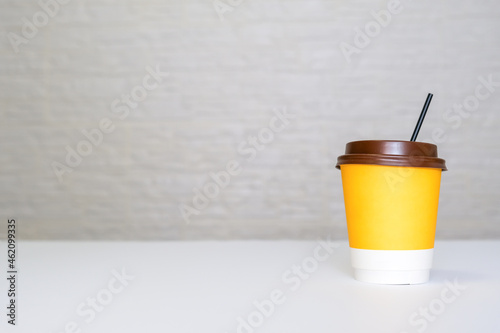 A yellow paper disposable takeout coffee cup with a brown cap and a straw on white background against light brick wall with copy space