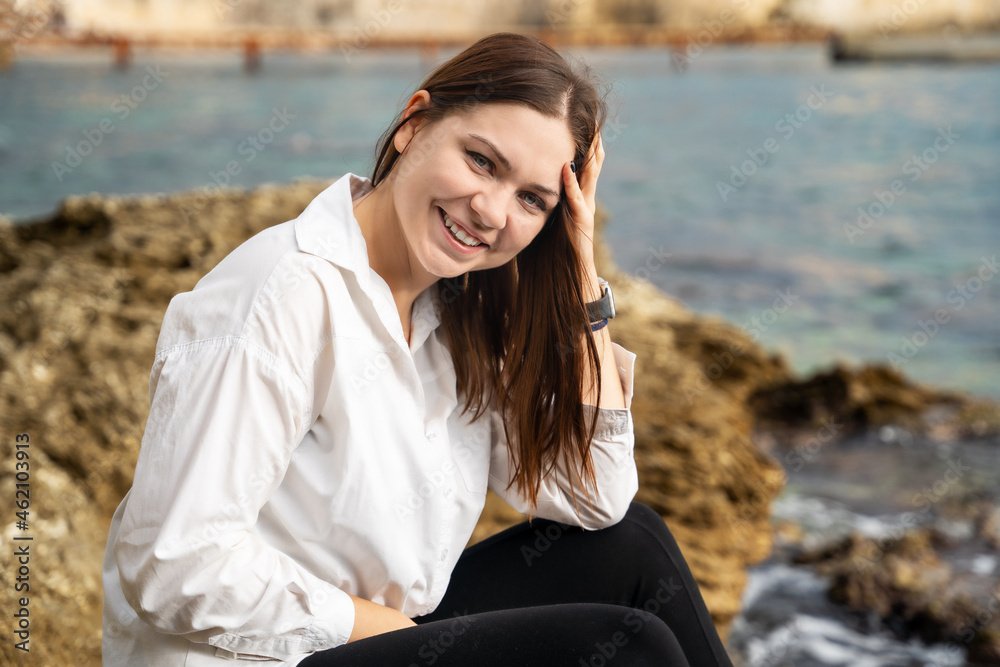 Attractive smiling brunette woman in white shirt sits on rocky seashore, walking in cloudy weather, blurred background. Outdoor activities during vacation. Posing female model. Attractive smiling brunette woman in white shirt sits on rocky seashore, walking in cloudy weather, blurred background. Outdoor activities during vacation. Posing female model.