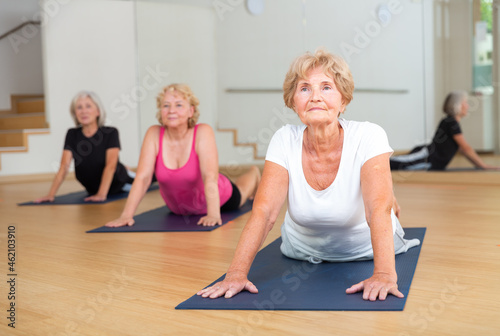 Group of aged ladies doing cobra pose on rugs in fitness room.