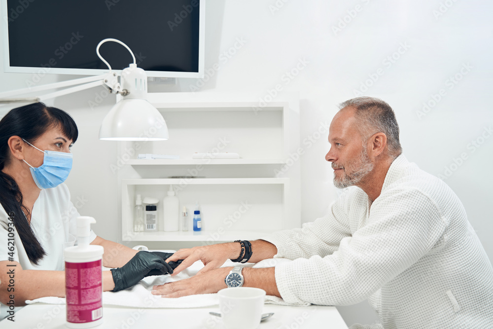 Fototapeta premium Manicurist in medical mask doing manicure for man in salon