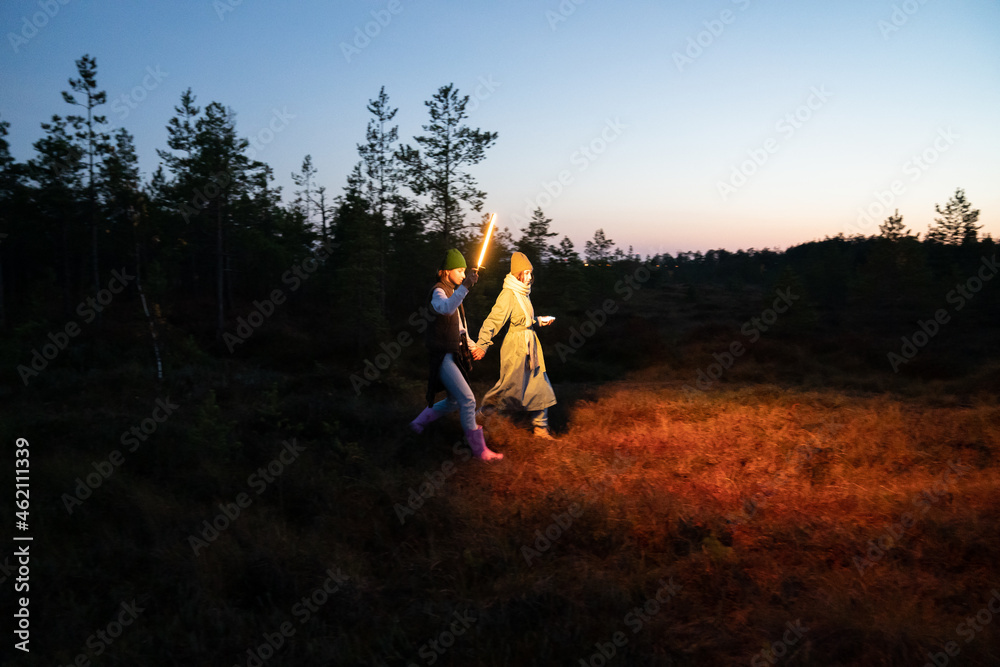 Two young women get lost in countryside after sunset walk holding hands highlighting trail or pathway with led lamp. Female friends returning from forest after hiking in autumn nature at night in dusk