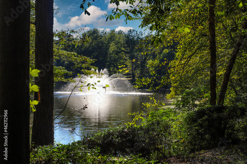 a gorgeous shot of a majestic water fountain in the middle of still lake water with lush green and autumn colored trees and plants reflecting off the lake water with blue sky and clouds at Lenox Park photo