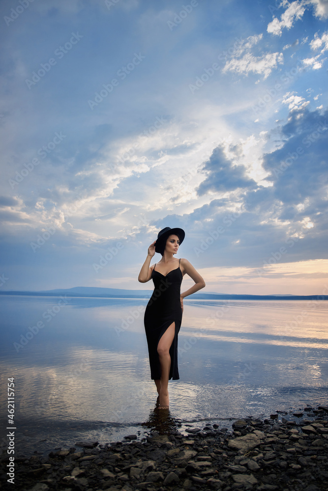Perfect brunette beauty woman in a black hat and a black dress poses near a lake against a blue sky. Long hair woman and beautiful beauty makeup on her face Perfect brunette beauty woman in a black hat and a black dress poses near a lake against a blue sky. Long hair woman and beautiful beauty makeup on her face