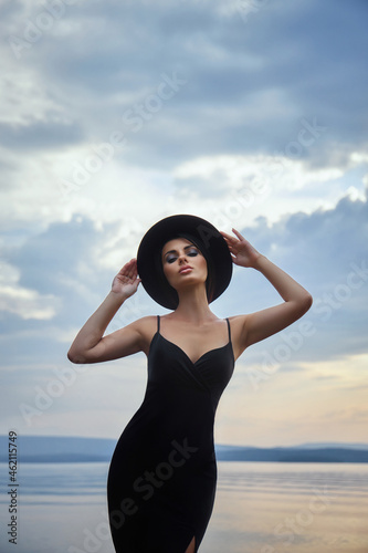 Perfect brunette beauty woman in a black hat and a black dress poses near a lake against a blue sky. Long hair woman and beautiful beauty makeup on her face