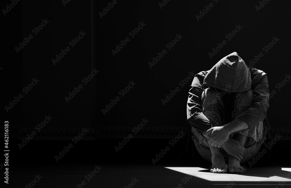 Light and shadow on surface of hopeless man in hoody sitting alone with hugging his knees on the floor in empty dark room in black and white style - obrazy, fototapety, plakaty 