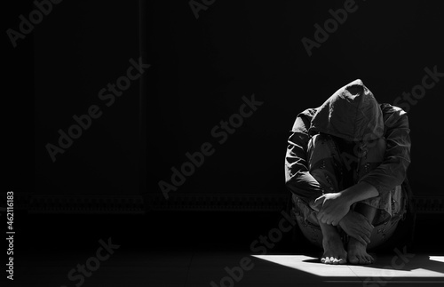 Light and shadow on surface of hopeless man in hoody sitting alone with hugging his knees on the floor in empty dark room in black and white style photo